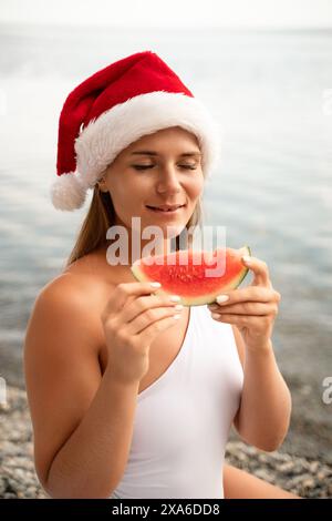 Une femme mange une pastèque tout en portant un chapeau de père noël rouge. La scène est légère et festive, car la femme profite d'une collation rafraîchissante sur un Banque D'Images