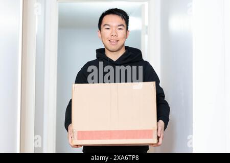 Un jeune homme tient une boîte en carton, emménageant dans un nouvel appartement ou une nouvelle maison. Il porte un sweat à capuche noir et sourit à la caméra Banque D'Images