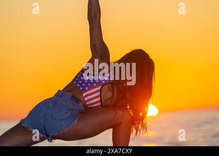 Une femme dans un haut de bikini pratique le yoga sur la plage, avec le soleil se couchant devant elle Banque D'Images