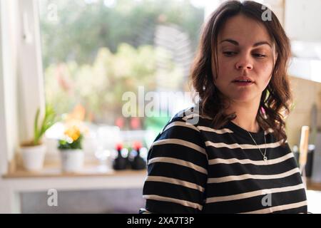 Une femme d'âge moyen aux cheveux foncés se tient dans un logsleeve rayé devant la fenêtre. Concept de vie familiale établie Banque D'Images