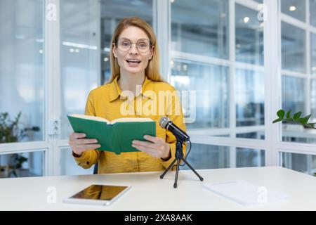 Femme en chemise jaune enregistrant podcast avec un livre et un microphone, assise dans un environnement de bureau moderne. Banque D'Images