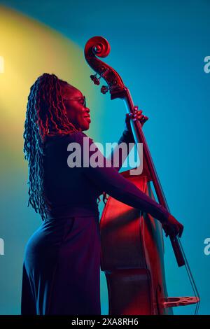 Jeune femme afro-américaine, musicienne jouant à la contrebasse dans la lumière néon sur fond dégradé coloré. Banque D'Images