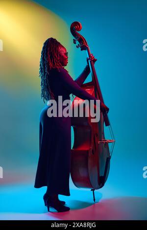 Jeune femme talentueuse afro-américaine, musicienne jouant à la contrebasse en néon sur fond dégradé coloré. Banque D'Images