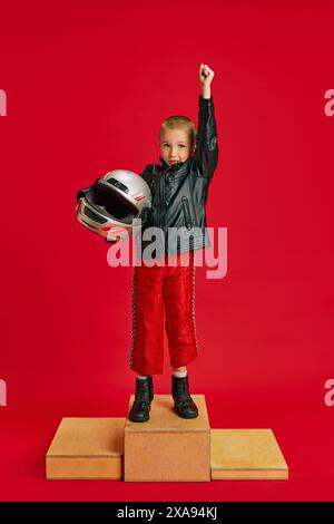 Enfant, petit garçon habillé en tenue de course se tient sur scène en bois et célébrant la victoire dans la compétition imaginaire sur fond rouge vif. Banque D'Images