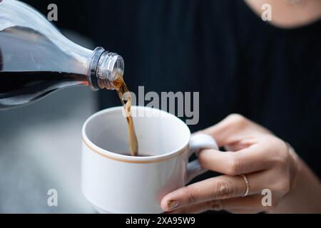 Gros plan d'une personne versant une boisson sombre d'une bouteille en plastique dans une tasse en céramique blanche. Banque D'Images