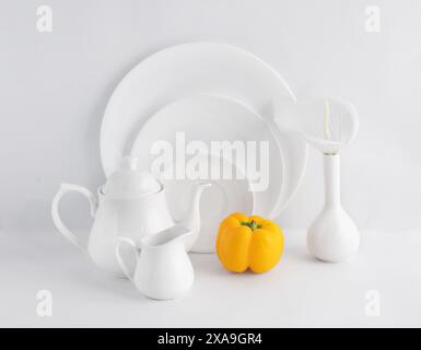 Vue rapprochée de la vaisselle blanche qui comprend une théière, pot à lait, trois assiettes blanches et un petit vase avec un poivron jaune. Banque D'Images
