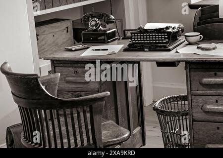 Bureau de l'ère technologique des années 1930 vintage avec bureau de style ancien, machine à écrire et téléphone de bureau à cadran rotatif noir rétro, avec chaise de bureau en bois et divers papiers de bureau etc de l'époque. Traitement sépia B&W 1920s/1930s Banque D'Images