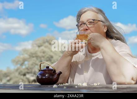 vue de face d'une femme buvant une tasse de thé à la camomille naturelle sur une table en bois dans un champ avec des fleurs de camomille autour d'elle. Banque D'Images