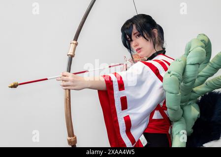 Une cosplayer féminine dans une tenue blanche et rouge et une cloche autour de son cou posant avec un arc et une flèche Banque D'Images