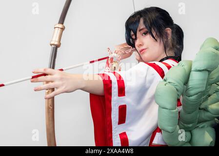 Une cosplayer féminine dans une tenue blanche et rouge et une cloche autour de son cou posant avec un arc et une flèche Banque D'Images