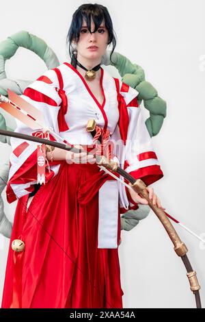 Une cosplayer féminine dans une tenue blanche et rouge et une cloche autour de son cou posant avec un arc et une flèche Banque D'Images