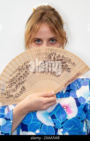 Une cosplayer féminine dans un kimono bleu avec une ceinture rouge tenant un ventilateur en bois devant son visage Banque D'Images