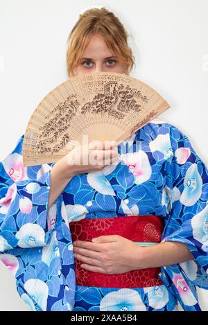 Une cosplayer féminine dans un kimono bleu avec une ceinture rouge tenant un ventilateur en bois devant son visage Banque D'Images