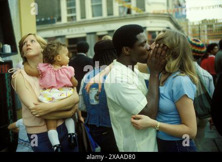 1970s UK jeune homme noir britannique embrasse son partenaire blanc britannique, derrière eux un ami tient leur bébé de race mixte enfant. Carnaval de Notting Hill, Londres, Angleterre 27 août 1979. HOMER SYKES Banque D'Images