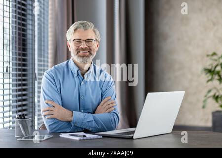 Homme d'affaires senior confiant portant des lunettes et une chemise bleue travaillant à son bureau avec un ordinateur portable dans un cadre de bureau moderne, souriant à la caméra. Banque D'Images