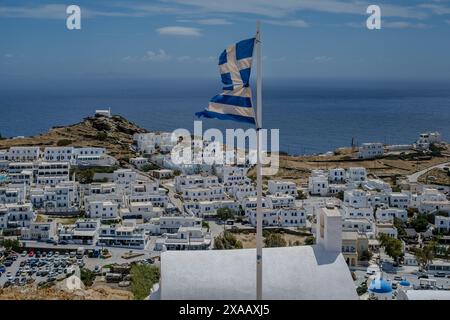 IOS, Grèce - 4 mai 2024 : vue panoramique d'un drapeau national grec, divers hôtels blanchis à la chaux, une église au sommet de la colline à iOS en Grèce Banque D'Images