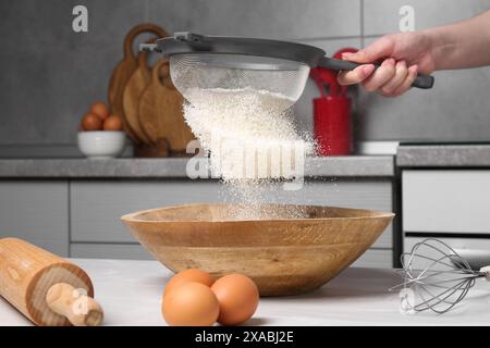 Femme tamisant la farine dans le bol à la table en bois blanc dans la cuisine, gros plan Banque D'Images