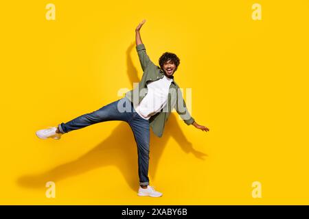 Photo de portrait du jeune mec mexicain drôle en chemise kaki et jeans debout garder l'équilibre isolé sur fond de couleur jaune Banque D'Images