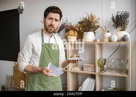 Homme mature barbu dans un tablier vert tenant une plaque cassée, debout près des étagères remplies de verrerie et d'articles décoratifs dans le studio de décoration. Banque D'Images