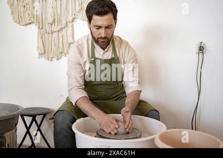 Potier masculin axé sur la sculpture de plats en céramique à l'aide d'une roue de potier. Homme mûr dans le tablier travaillant dans le studio de poterie. Banque D'Images