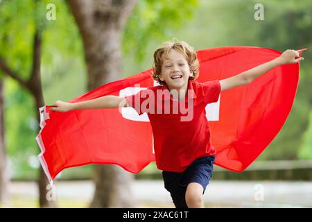 Enfant qui court avec le drapeau suisse. Petit enfant suisse célébrant la fête nationale. Fan suisse sur le terrain de football en regardant l'équipe de football jouer. Banque D'Images