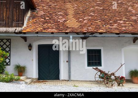 Photographie d'une ancienne ferme alpine traditionnelle avec des murs blancs, un toit en tuiles rouges, des cadres de fenêtres vertes et des portes avec du matériel agricole ancien à proximité Banque D'Images