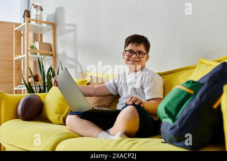 Un adorable garçon avec le syndrome de Down assis sur un canapé jaune, utilisant un ordinateur portable. Banque D'Images