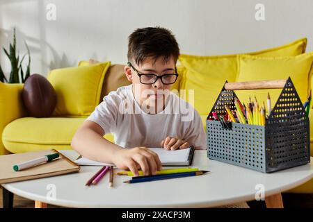 Un petit enfant atteint du syndrome de Down est assis à une table, engrossé dans l'écriture dans son cahier avec des crayons de couleur. Banque D'Images