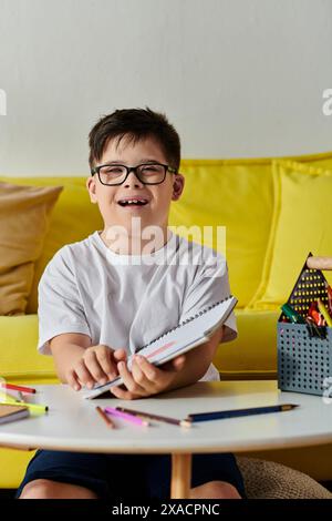 Enfant adorable avec le syndrome de Down avec des lunettes à table, coloration dans un cahier avec des crayons de couleur. Banque D'Images