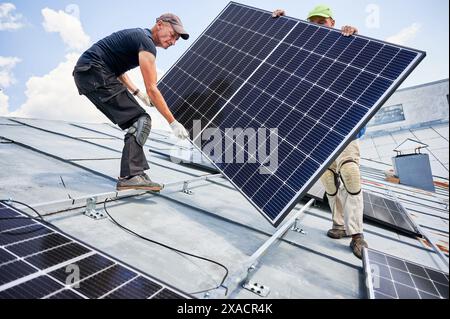 Ouvriers construisant le système de panneau solaire sur le toit métallique de la maison. Deux hommes installateurs portant le module solaire photovoltaïque à l'extérieur. Concept de production d'énergie alternative, verte et renouvelable. Banque D'Images