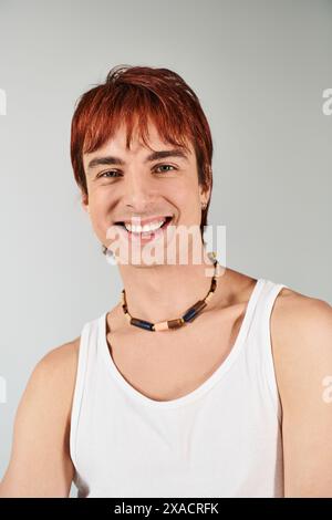 Un jeune homme élégant aux cheveux rouges frappant une pose dans un débardeur blanc sur un fond de studio gris. Banque D'Images