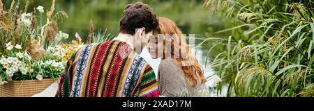Un homme et une femme, vêtus d'une tenue de style boho, blottis ensemble sous une couverture confortable dans un parc verdoyant luxuriant. Banque D'Images
