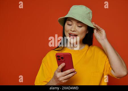 Une femme asiatique à la mode dans un chapeau tenant un téléphone portable sur un fond orange. Banque D'Images