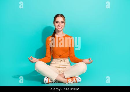 Photo pleine longueur de femme satisfaite porter pantalon beige crop top assis sur le sol dans la pose de méditation isolé sur fond de couleur turquoise Banque D'Images