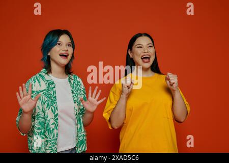 Deux femmes d'origine asiatique se tiennent debout avec les mains levées dans un cadre de studio sur un fond orange. Banque D'Images