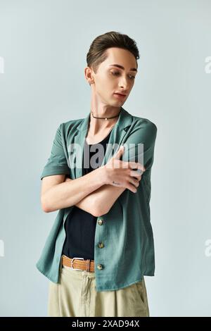 Une jeune personne queer pose en toute confiance dans un studio portant une chemise verte et un pantalon bronzé, exprimant la fierté LGBT sur un fond gris. Banque D'Images