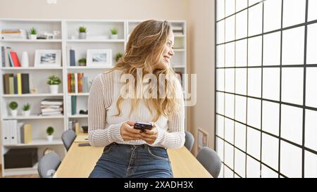 Jeune et belle femme d'affaires hispanique rayonne de confiance tout en souriant et en gérant habilement son travail en ligne à l'aide de son smartphone dans l'elegan Banque D'Images
