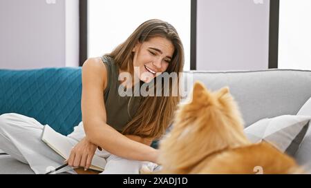 Belle jeune femme hispanique aime s'asseoir avec son chien joyeux à la maison, rayonnant de positivité et de confiance de son sourire joyeux dans le vivant R Banque D'Images