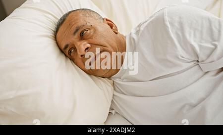 Un homme hispanique d'âge moyen semble contemplatif tout en se reposant dans une chambre avec une literie blanche. Banque D'Images