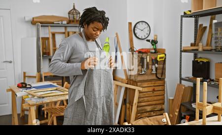 Femme afro-américaine avec des dreadlocks portant un tablier dans un atelier de menuiserie examinant un textile. Banque D'Images