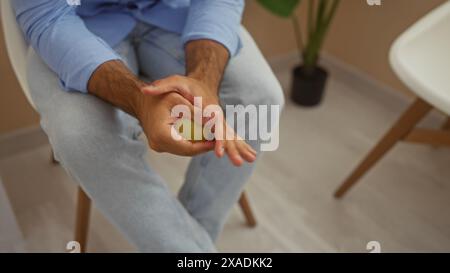 Jeune homme hispanique assis à l'intérieur dans une salle d'attente, pressant une balle de stress avec ses mains dans une posture détendue. Banque D'Images