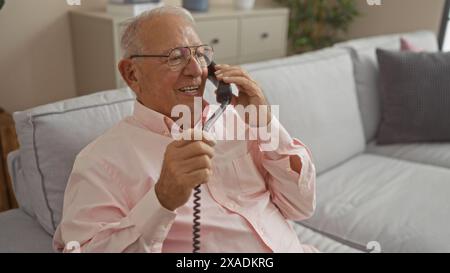 Homme caucasien senior aux cheveux gris riant au téléphone tout en étant assis sur un canapé dans un salon confortable. Banque D'Images