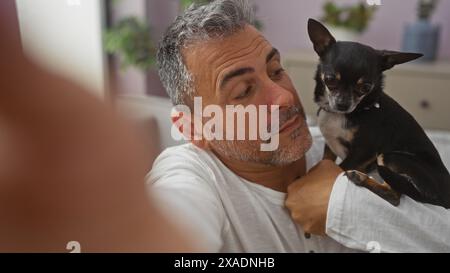 Homme hispanique d'âge moyen tenant chihuahua à l'intérieur à la maison, regardant le petit chien dans un salon d'appartement confortable. Banque D'Images