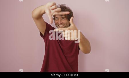Un jeune homme hispanique joyeux avec une barbe faisant un cadre avec ses mains sur un fond de mur rose. Banque D'Images