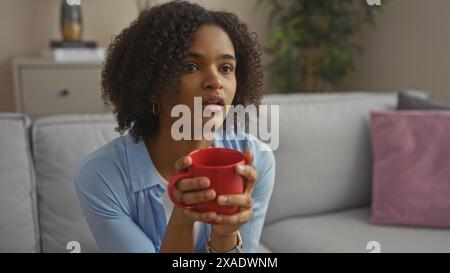 Une belle femme afro-américaine aux cheveux bouclés tient une tasse rouge tout en étant assise sur un canapé dans un salon confortable. Banque D'Images