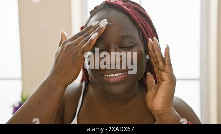 Une femme souriante avec des tresses applique de la crème sur son visage à l'intérieur, dépeignant un moment de routine beauté. Banque D'Images