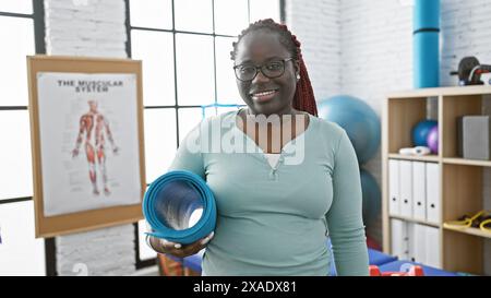 Une femme afro-américaine souriante avec des tresses tient des tapis de yoga dans la salle de réadaptation d'une clinique de physiothérapie, mettant en valeur le bien-être et la récupération. Banque D'Images