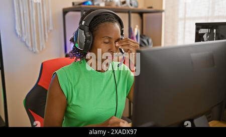 Une femme afro-américaine portant des écouteurs travaille intensément à un ordinateur dans un bureau à domicile moderne. Banque D'Images