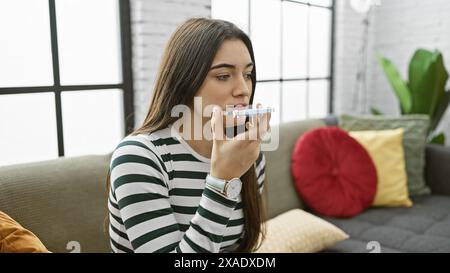 Jeune femme hispanique parle sur smartphone dans un salon confortable bien éclairé, évoquant la communication décontractée à la maison. Banque D'Images