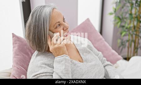 Une femme âgée discute sur un smartphone tout en s'inclinant confortablement dans sa chambre moderne Banque D'Images